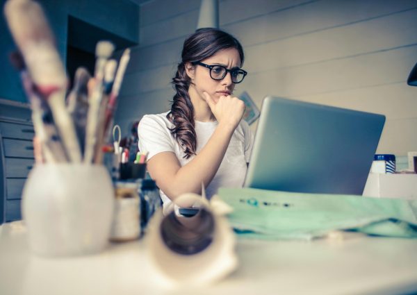 Young woman with glasses deeply focused on a laptop surrounded by art supplies in a home office.